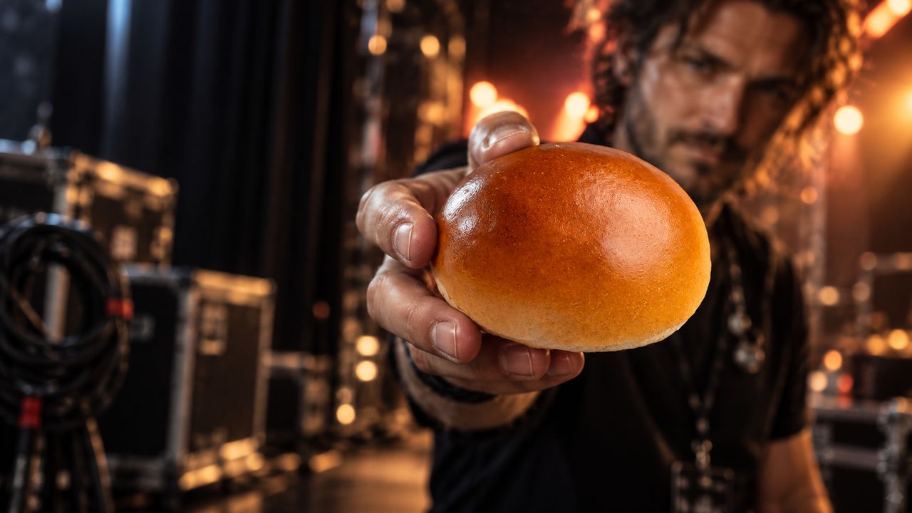 A hand holding a golden bun towards the camera in a dim backstage area with warm stage lights behind